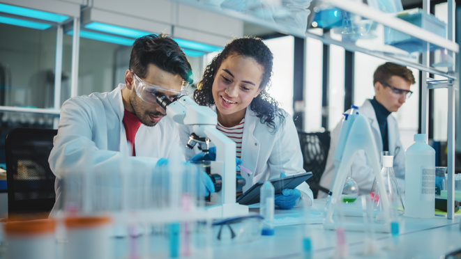 Zwei junge Wissenschaftlern, die ein Mikroskop und ein digitales Tablet verwenden, Probenanalysen durchführen und sprechen. 