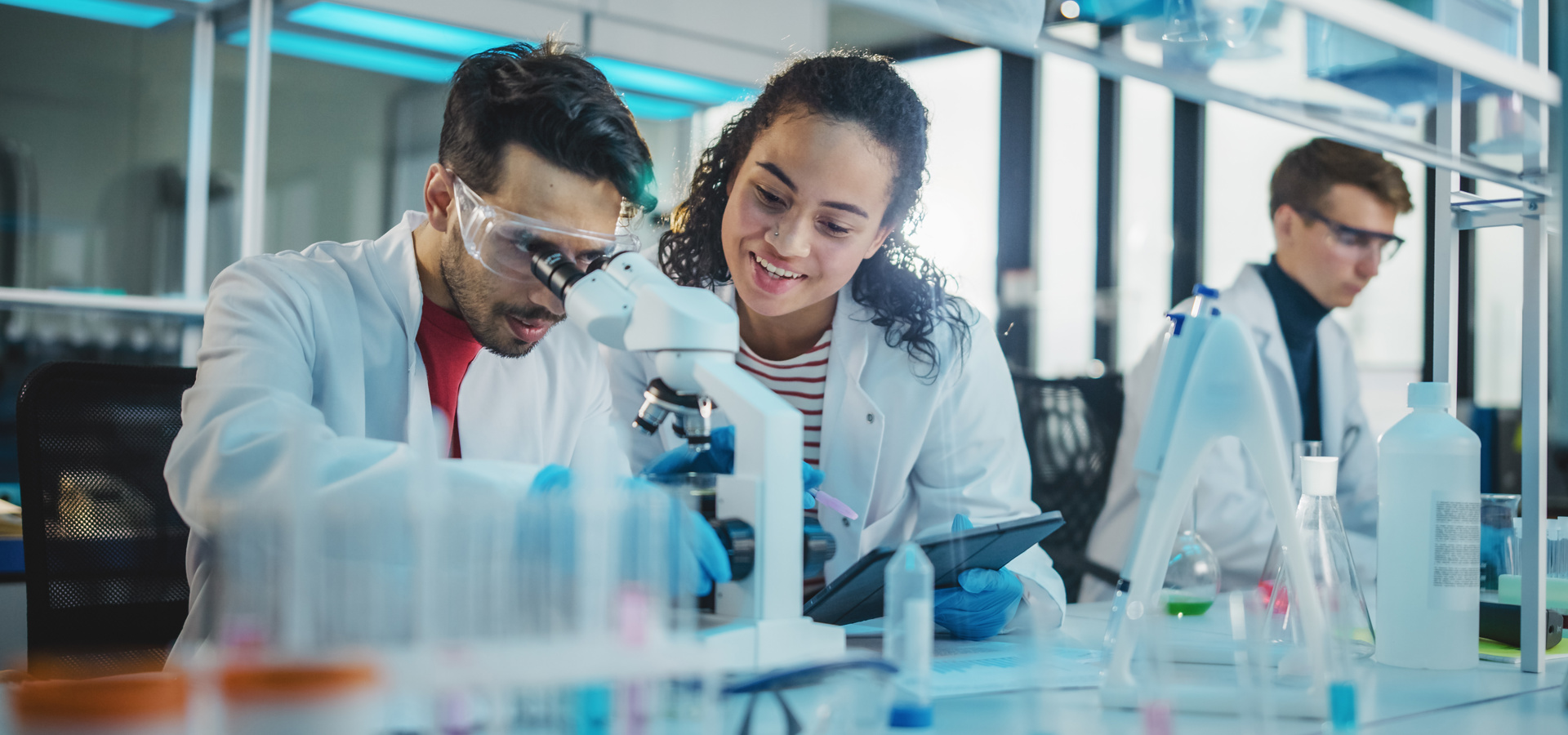 Zwei junge Wissenschaftlern, die ein Mikroskop und ein digitales Tablet verwenden, Probenanalysen durchführen und sprechen.